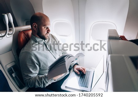 Smart casual male book editor holding literature and making notes in laptop application for creating text during time in airplane before business meeting with writer, journalist working during flight