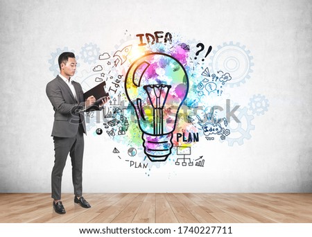 Serious and successful young Asian businessman writing on clipboard in concrete wall room with colorful business idea sketch. Concept of creativity and business strategy