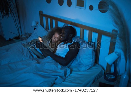 High angle portrait of young mixed-race couple lying in bed at night looking at smartphone screen while talking, copy space