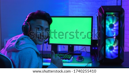 e cyber sport gamer has thumb up gesture and smile to you in front of green screen monitor with powerful personal computer on the table at home