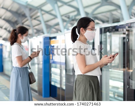 Two Asian women wearing medical face mask, using smartphone waiting for metro at train station platform, standing distance apart from other people. New normal trend and social distancing concept Royalty-Free Stock Photo #1740271463