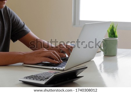 An Asian man uses a laptop at home while sitting at a wooden table. Man hands typing on a notebook keyboard. The concept of young people working at mobile devices.