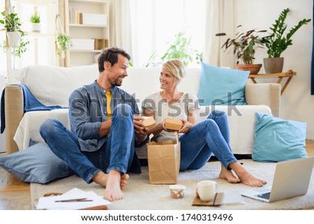 Happy couple sitting on floor indoors at home, eating hamburgers. Royalty-Free Stock Photo #1740319496