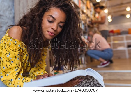 Portrait of young female student sitting at library reading a book with with her friends in background.