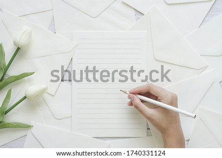 Female hand writing letter on white wooden desk background with white tulips. Top view