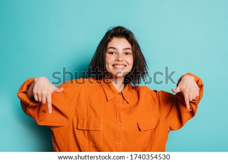 Picture of positive cheerful young woman or teenager isolated over blue background. Happy girl point down with index fingers. Stylish modern woman wear orange shirt