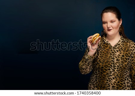 Fat woman in a leopard print blouse with a hamburger in her hand. Empty space on the left. Blue background.