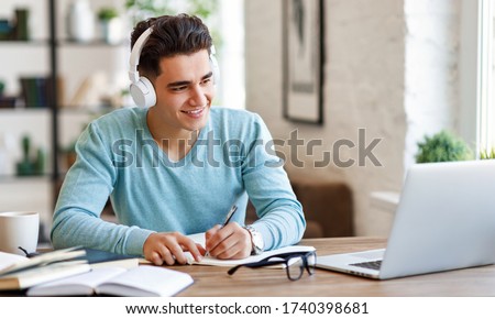 Happy ethnic guy in headphones smiling and writing in notebook while sitting at table and listening to teacher during online lecture at home
