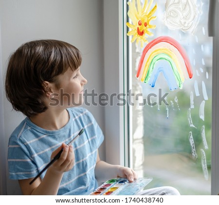 A little boy draws a rainbow on a window during a coronavirus pandemic.