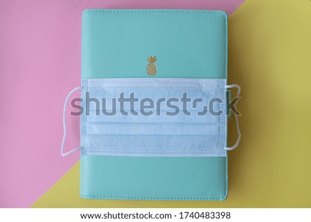Blue turquoise notebook ,notepad , planner  with a gold pineapple   And blue protective medical mask on a top from coronavirus disease on yellow and pink background .