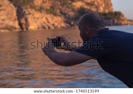 A man taking photos at sunset in Cala Llamp with his mobile phone. Beach cove in Mallorca