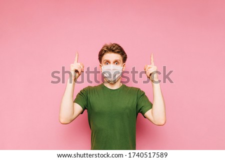 Shocked young man in gauze medical mask isolated on pink background, looking at camera and showing fingers up on copy space. Coronavirus pandemic. Quarantine. covid-19.