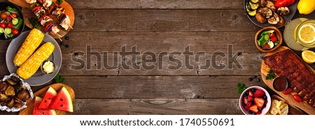 Summer BBQ or picnic food double side border over a rustic wood banner background. Various grilled meats, vegetables, fruits, salad and potatoes. Above view with copy space.