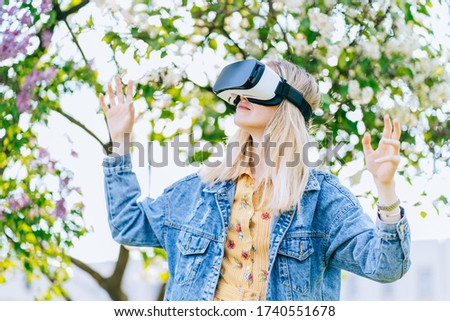 Blond romantic woman wearing virtual reality goggles outside in spring nature. Hipster girl using VR glasses walking in green garden, modern technology concept.