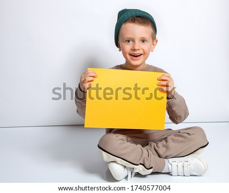 A handsome European boy with blue eyes holds an orange sheet of paper, sits on a white background and smiles. Place for text, banner, surface for inscriptions and advertising.