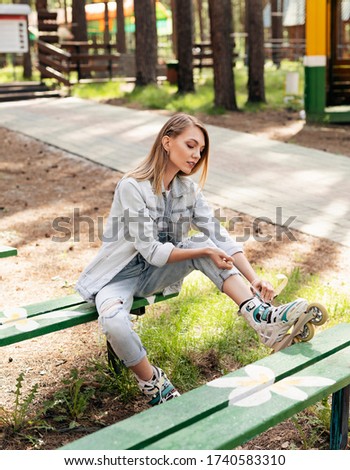 Portrait of a beautiful young blonde girl who pulls up roller skates, sitting on a bench in a city Park. Hot summer day. Girl on roller skates in the fresh air. A girl in a denim suit on a bench.