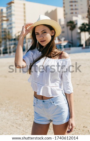 girl in a stylized country costume cowboy stands on the beach sand against the background of high-rise buildings holding a hat with her hand and looking carefully at the camera