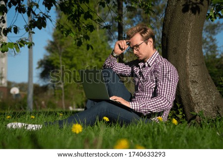 A young Caucasian man sits under a tree, typing on a laptop. Internet concept. Freelancer working in the park on the grass.