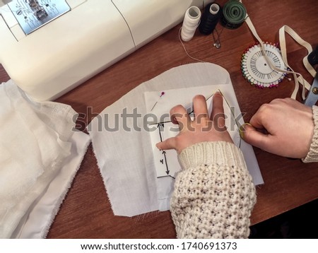 Seamstress outlines a paper pattern with yellow chalk on a white cloth. Female hands draw a pattern of a medical mask on the background of a sewing machine and accessories, background with copy space