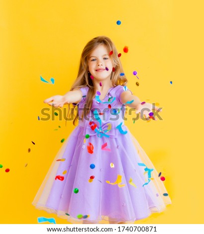 happy little girl with blond hair and in a blue dress catches confetti on a yellow background, holiday concept