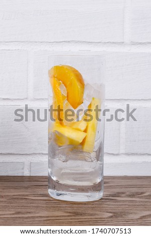 Tall glass with ingredients for freshly squeezed fruit juice, before pressing.
Pineapple, mango and orange and ice on a wooden table by a white brick wall. Healthy smoothie. 