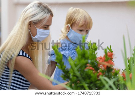 Mom wears her daughter a medical mask. Outdoors, against a wall and flowers, close-up.