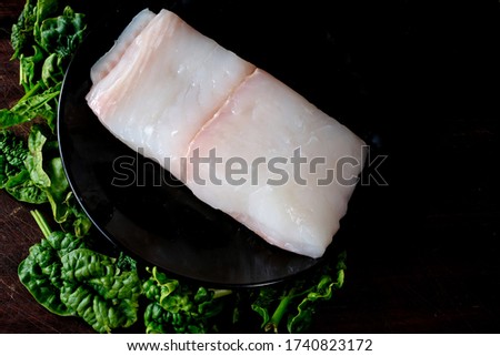Top view of beautiful piece of fresh raw halibut fish on black plate with green spinach at side. Copy space