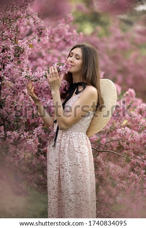 Portrait of a young woman in a blooming garden.
She gently sniffs a twig with pink flowers. Image with selective focus and toning. Focus on the girls.