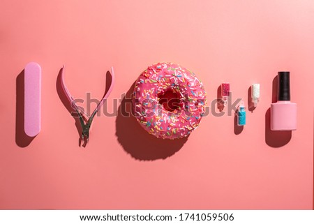 On a pink background are tools, beads for design, nail polish and a appetizing beautiful glazed donut. Concept of summer mood, bright colors and tasty sweet and trendy manicure and pedicure. Top view