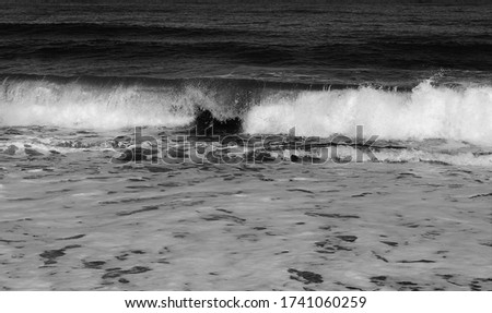 Monochrome picture of waves spray as water hits submerged rocks at the edge of the sand.