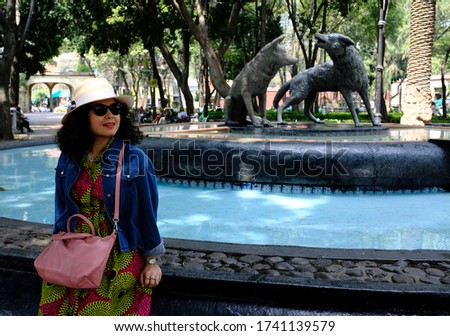 An Asian female tourist posing for a picture in front of the famous drinking coyotes volcanic stone statue in Coyoacán neighbourhood, Mexico City, Mexico.