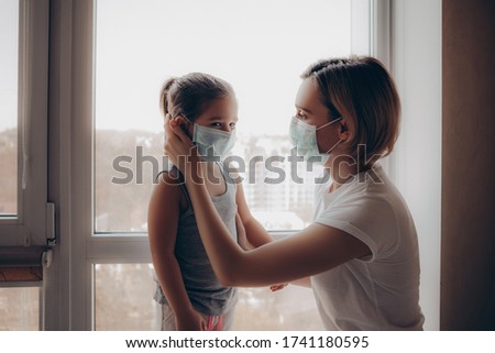 Family Mom and Daughter in Medical Mask. Young Woman and Child little girl sitting by the Window in Protective Masks against the Virus. Mom carefully corrects the mask on the face of the  child        Royalty-Free Stock Photo #1741180595