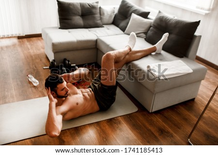 Young man doing abs exercising at home on the couch. Cut view of hardwoking t-shirtless guy sportsman in workout activity at his apartment. dumbbells on pictures lying on floor. Trying to get better.