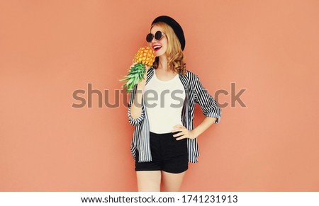 Portrait of happy young smiling woman singing with pineapple wearing a black hat, sunglasses, striped shirt over wall background
