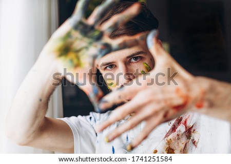 The artist creates a frame with his hands soiled with watercolor paints. Portrait of a young creative artist, the process of creating a picture. Royalty-Free Stock Photo #1741251584