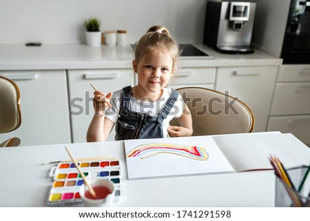 A girl child of 4 to 7 years old sits in the house and paints on paper. The child draws a rainbow, focused on the picture.  