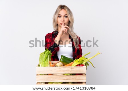 Farmer girl holding a basket full of fresh vegetables over isolated white background showing a sign of silence gesture putting finger in mouth