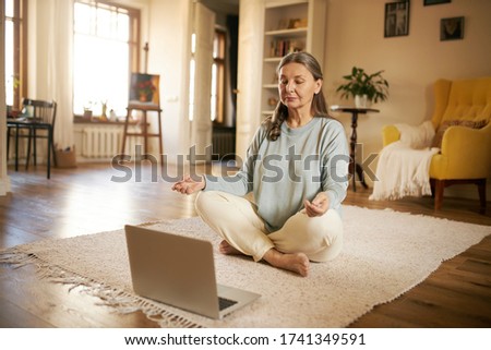 Meditation, self-awareness, healthy body and mind. Casually dressed mature woman sitting on floor in front of laptop, legs crossed, closing eyes, meditating, listening to calm music or affirmations Royalty-Free Stock Photo #1741349591