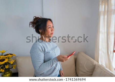 A picture of a woman standing near the window while taking a call on her cell phone in the living room of her house.