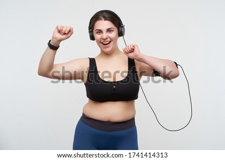 Glad young cute brunette chubby female with casual hairstyle dancing with raised hand and smiling happily while listening to music with earphones, posing over white background