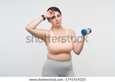 Exhausted young dark haired plump woman with blue dumbbells wiping sweat while being tired after hard training, posing over white background. Body positive concept