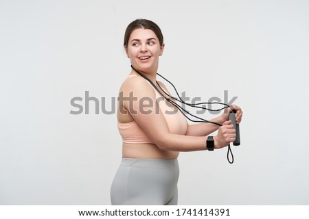 Positive young lovely dark haired chubby lady dressed in sports bra and leggins wearing jumping-rope on her neck and looking cheerfully over her shoulder. Body positive concept