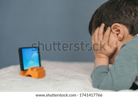 little boy touching head while watching educational cartoon on smartphone while lying on bed