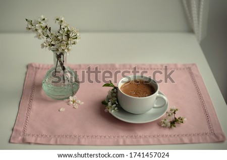 A Cup of coffee and a bouquet of white flowers in a transparent vase on the table . Pink tissue paper. Pink tablecloth