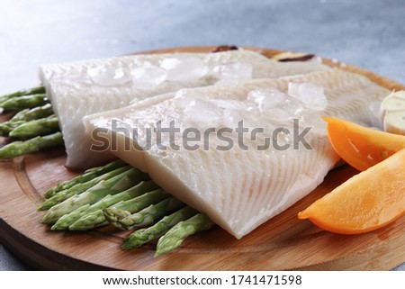 Seafood. Raw white fish fillet, halibut fillet with asparagus, ice, tomatoes, garlic, salt on a wooden board on a light grey background. Background image, copy space 