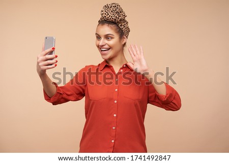 Happy young lovely brown haired female with headband smiling cheerfully at camera of her phone and raisinghand in hello gesture while having video call, posing over beige background