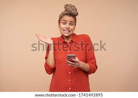 Bewildered young green-eyed brown haired lady grimacing her face and raising perplexedly palm while looking confusedly at camera, standing over beige background