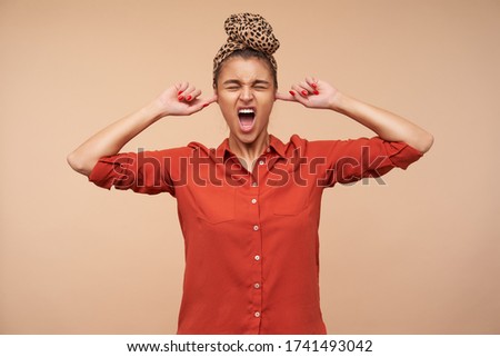 Indoor photo of young brunette woman with headband keeping her eyes closed while grimacing face and inserting forefingers into ears, isolated over beige background