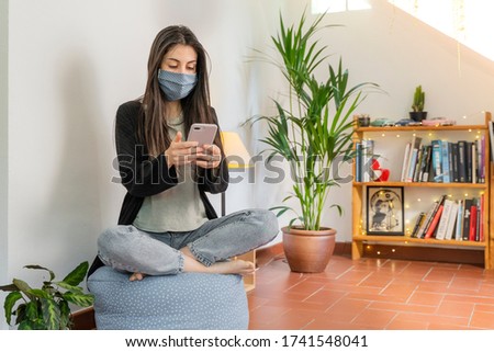 Coronavirus. Woman sitting down using mobile phone. Wearing mask protection and recovery from the illness in home. Quarantine. Patient isolated to prevent infection. Pandemia. Home.