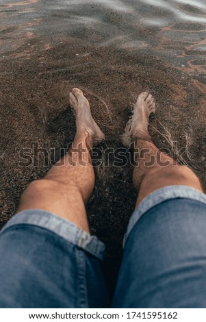 Stock photo of a man in shorts sitting on the seashore at the beach with his feet in the water